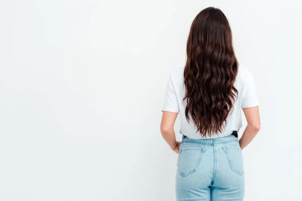 Gehakt beeld. Achteraanzicht van jonge vrouw, gekleed in een wit T-shirt en blauwe jeans, staat voor lichte achtergrond met zijn hand in broekzak. Maak je niet druk. Ruimte voor tekst, reclame. — Stockfoto