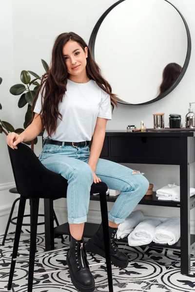 Menina encantadora em uma camiseta branca e jeans senta-se em uma cadeira perto de uma mesa de cabeleireiro, em um salão de beleza — Fotografia de Stock