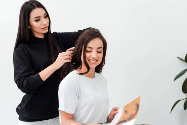 Smiling, attractive brunette is reading a magazine while her hairdresser combs her beautiful hair — 스톡 사진