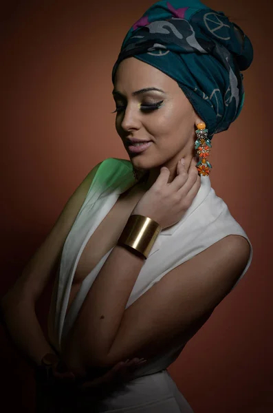 Beautiful woman with turban. Young attractive female with turban and golden accessories. Beauty fashionable woman with hair wrapped in turban. Pretty Caucasian model wearing earrings posing in studio. — Stock Photo, Image