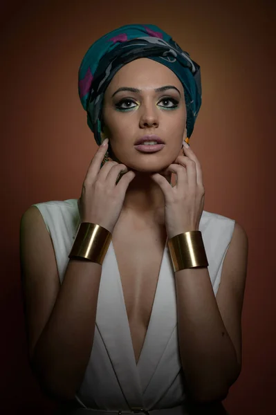 Hermosa mujer con turbante. Joven atractiva hembra con turbante y accesorios dorados. Belleza mujer de moda con el pelo envuelto en turbante. Bonita modelo caucásica con pendientes posando en estudio . —  Fotos de Stock