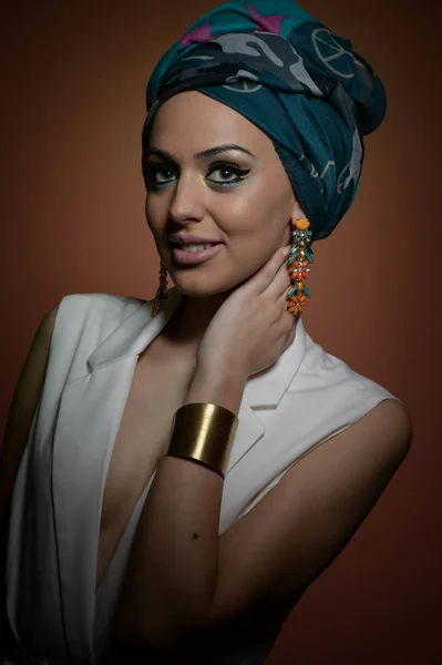 Vacker kvinna med turban. Ung attraktiv kvinna med turban och gyllene accessoarer. Skönhet fashionabla kvinna med hår insvept i turban. Ganska kaukasiska modell bär örhängen poserar i studio. — Stockfoto