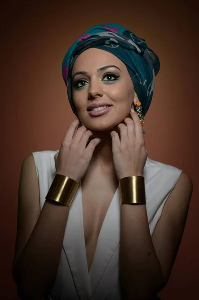 Beautiful woman with turban. Young attractive female with turban and golden accessories. Beauty fashionable woman with hair wrapped in turban. Pretty Caucasian model wearing earrings posing in studio. — Stock Photo, Image