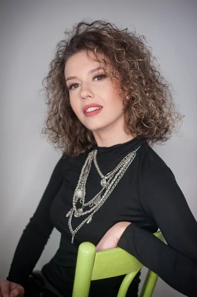 Retrato de un modelo de moda impresionante sentado en una silla en el estudio. De cerca Retrato de mujer hermosa joven. Retrato de mujer atractiva sentada en una silla verde aislada sobre fondo gris —  Fotos de Stock