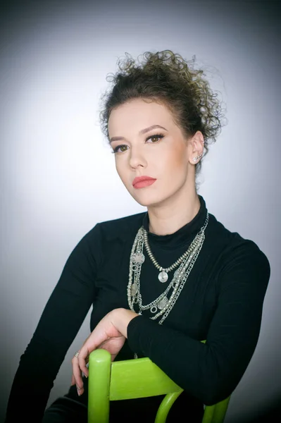 Portrait of a stunning fashionable model sitting in a green chair, studio shot. Close up portrait of young beautiful woman with curly hair. Attractive woman with black turtle neck blouse isolated on grey background — Stock Photo, Image