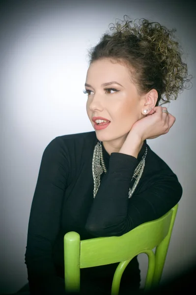 Portrait of a stunning fashionable model sitting in a green chair, studio shot. Close up portrait of young beautiful woman with curly hair. Attractive woman with black turtle neck blouse isolated on grey background — Stock Photo, Image