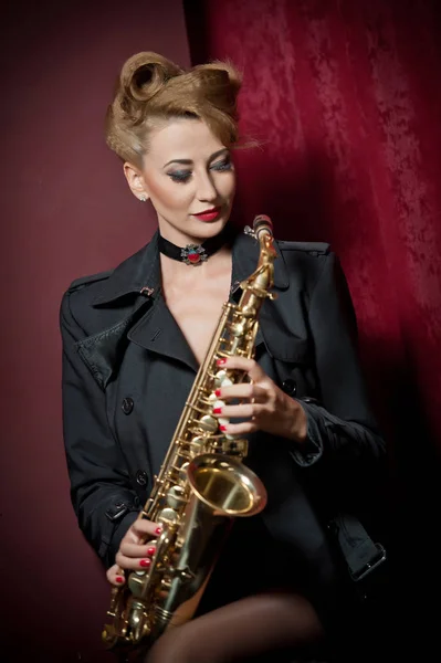 Sexy mujer atractiva con saxofón posando sobre fondo rojo. Joven rubia sensual jugando al saxo. Instrumento musical, jazz. Hermosa mujer en negro posando con saxofón, toma de estudio —  Fotos de Stock