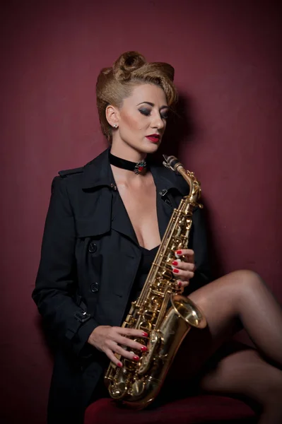 Sexy mujer atractiva con saxofón posando sobre fondo rojo. Joven rubia sensual jugando al saxo. Instrumento musical, jazz. Hermosa mujer en negro posando con saxofón, toma de estudio — Foto de Stock