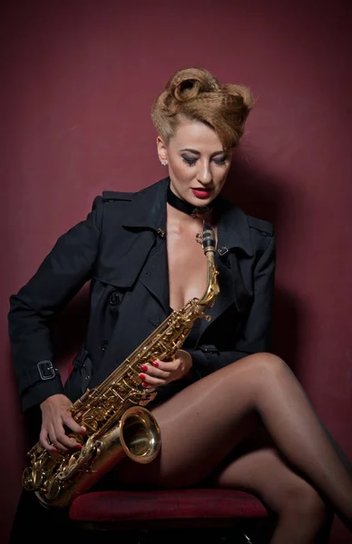 Sexy mujer atractiva con saxofón posando sobre fondo rojo. Joven rubia sensual jugando al saxo. Instrumento musical, jazz. Hermosa mujer en negro posando con saxofón, toma de estudio — Foto de Stock