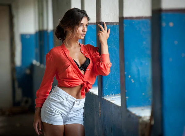 Beautiful girl with red shirt and white shorts posing in old hall with columns blue painted. Attractive long hair brunette, side view against ancient pillars. Dark hair young woman with gorgeous eyes — Stock Photo, Image