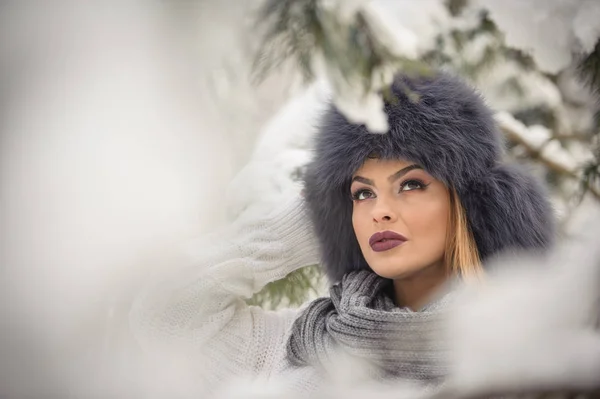 Bella donna in pullover bianco con berretto di pelliccia over-size godendo del paesaggio invernale nella foresta. Ragazza bionda posa sotto rami di alberi innevati.Attraente giovane femmina in giornata fredda brillante, trucco — Foto Stock