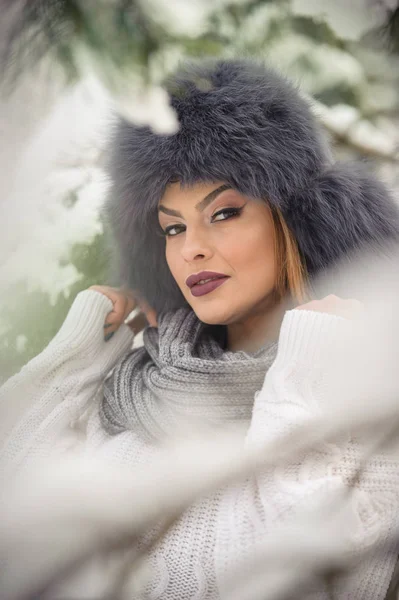 Bella donna in pullover bianco con berretto di pelliccia over-size godendo del paesaggio invernale nella foresta. Ragazza bionda posa sotto rami di alberi innevati.Attraente giovane femmina in giornata fredda brillante, trucco — Foto Stock