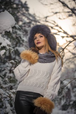 Ormandaki kış manzarası zevk aşırı büyüklükteki kürk kapaklı beyaz kazak güzel kadın. Sarışın kız karla kaplı ağaçların dalları altında poz. Çekici genç bayan parlak soğuk günde, makyaj