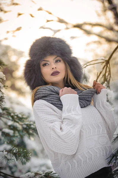 Schöne Frau in weißem Pullover mit übergroßer Pelzmütze, die die Winterlandschaft im Wald genießt. blondes Mädchen posiert unter schneebedeckten Bäumen verzweigt.attraktive junge Frau in strahlend kaltem Tag, Make-up — Stockfoto