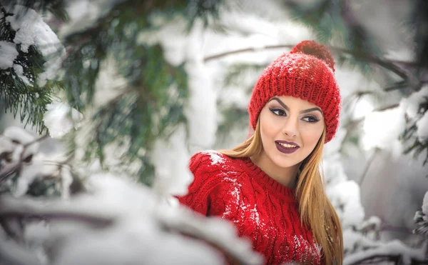 Женщина в красном с коричневым меховым плащом наслаждается зимними пейзажами в лесу. Блондинка позирует под заснеженными ветвями деревьев. Молодая женщина со снежинками вокруг в яркий холодный день, макияж — стоковое фото