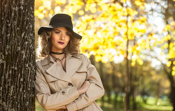 Piękna Blondynka kobieta krem płaszcz, długie nogi i czarny kapelusz w jesiennej scenerii. Portret przepiękny młody Elegant i zmysłowe kobiety z kręconymi włosami pozowanie w jesienny park. — Zdjęcie stockowe