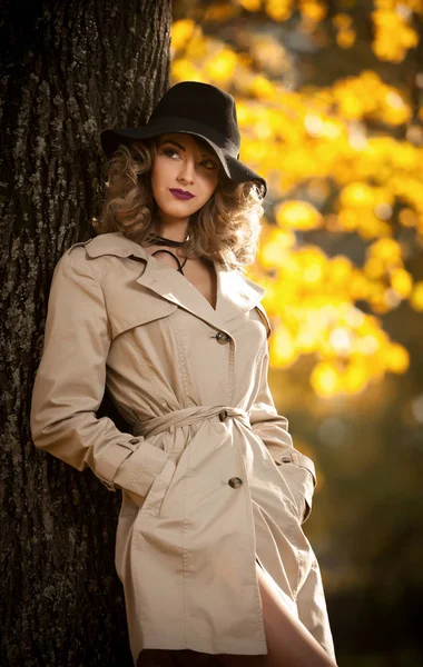Bela mulher loira com casaco de creme, pernas longas e chapéu preto em uma cena de outono. Retrato de uma jovem muito bonita Mulher elegante e sensual com cabelo encaracolado posando no parque de outono . — Fotografia de Stock