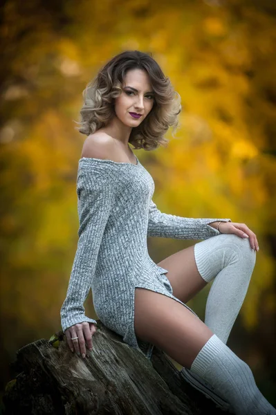 Sensual girl with long legs sitting on a stump in an autumnal scene.Long legs attractive blonde with curly hair relaxing in autumnal park.Fashionable young woman posing on a stump in the forest. — Stock Photo, Image