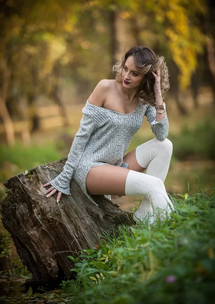 Sensuell tjej med långa ben sitter på en stubbe i en höstlig scen. Långa ben attraktiv blondin med lockigt hår avkopplande i höstlig park. Fashionabla ung kvinna poserar på en stubbe i skogen. — Stockfoto