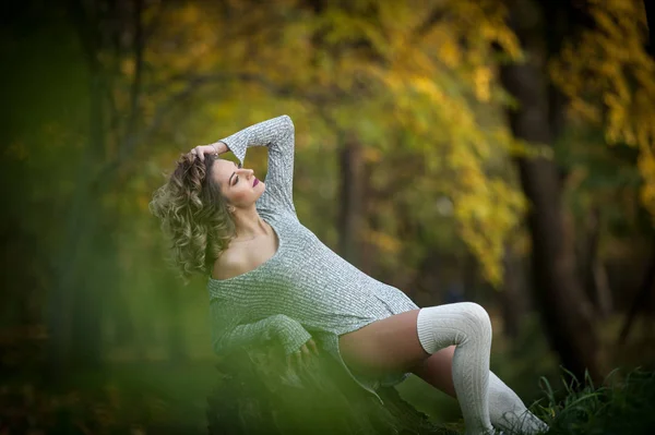 Sensuell tjej med långa ben sitter på en stubbe i en höstlig scen. Långa ben attraktiv blondin med lockigt hår avkopplande i höstlig park. Fashionabla ung kvinna poserar på en stubbe i skogen. — Stockfoto