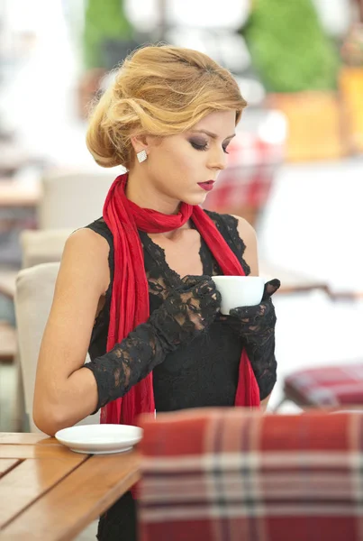 Señora atractiva de moda con pequeño vestido negro y bufanda roja sentado en la silla en el restaurante y beber café. Mujer rubia de pelo corto con maquillaje y corte de pelo creativo sosteniendo una taza de café —  Fotos de Stock
