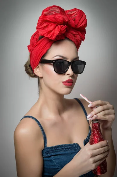 Mujer con turbante rojo y gafas de sol bebiendo de una botella con una pajita. Atractivo retrato de chica sosteniendo una botella, estudio filmado sobre fondo gris. Feliz joven mujer, concepto de publicidad —  Fotos de Stock