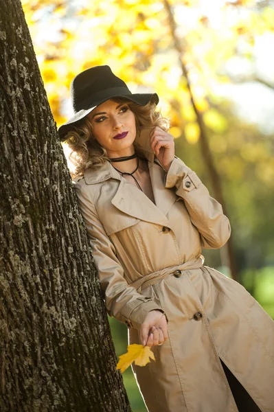 Schöne blonde Frau mit cremefarbenem Mantel, langen Beinen und schwarzem Hut in einer Herbstszene. Porträt einer sehr schönen jungen, eleganten und sinnlichen Frau mit lockigem Haar, die im Herbstpark posiert. — Stockfoto