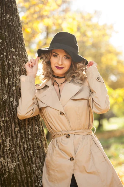 Bela mulher loira com casaco de creme, pernas longas e chapéu preto em uma cena de outono. Retrato de uma jovem muito bonita Mulher elegante e sensual com cabelo encaracolado posando no parque de outono . — Fotografia de Stock