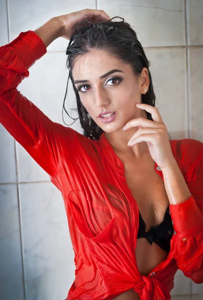 完璧なボディの濡れた髪と透明な赤いシャツ、完璧なぽっちゃりボディのシャワーの下で官能的なポーズでポーズをとるかわいいブルネットの女性。濡れたシャツでうれしそうな少女の肖像画 — ストック写真