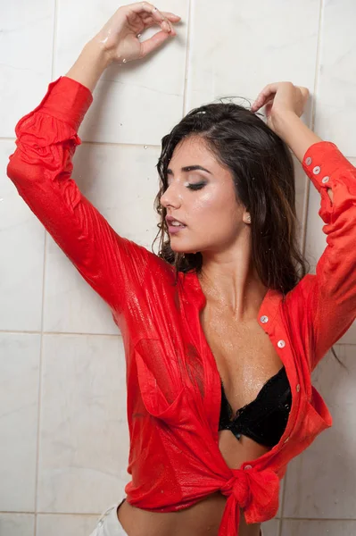 Mulher morena bonito com corpo perfeito posando em pose erótica sob o chuveiro com cabelo molhado e camisa vermelha transparente, corpo voluptuoso perfeito.Retrato de uma menina alegre na camisa molhada — Fotografia de Stock