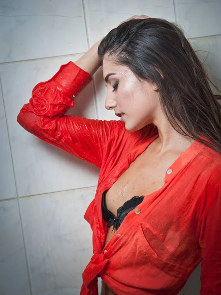 Mulher morena bonito com corpo perfeito posando em pose erótica sob o chuveiro com cabelo molhado e camisa vermelha transparente, corpo voluptuoso perfeito.Retrato de uma menina alegre na camisa molhada — Fotografia de Stock