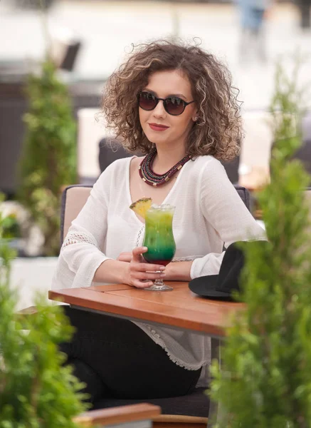 Gelukkig Brunette meisje met zonnebril zitten in het Park, het drinken van een glas koude groene SAP terwijl glimlachend in de afstand. Jonge mooie vrouw op de Bank drinken sap dragen een wit overhemd — Stockfoto