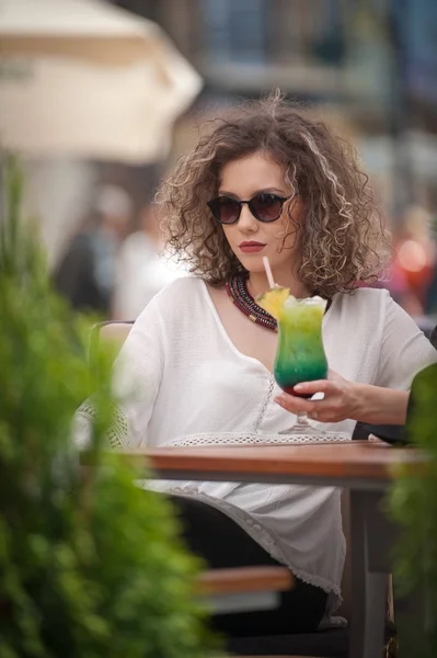 Gelukkig Brunette meisje met zonnebril zitten in het Park, het drinken van een glas koude groene SAP terwijl glimlachend in de afstand. Jonge mooie vrouw op de Bank drinken sap dragen een wit overhemd — Stockfoto