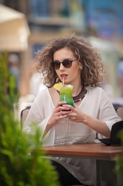 Gelukkig Brunette meisje met zonnebril zitten in het Park, het drinken van een glas koude groene SAP terwijl glimlachend in de afstand. Jonge mooie vrouw op de Bank drinken sap dragen een wit overhemd — Stockfoto