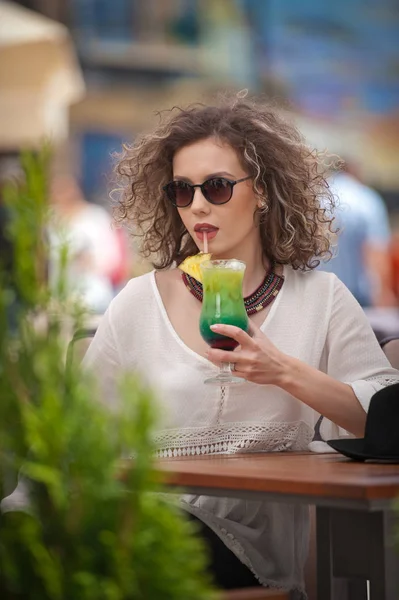 Happy Brunette Girl com óculos de sol Sentado no parque, bebendo um copo de suco verde frio enquanto sorri para a distância. Jovem mulher bonita no banco bebendo suco vestindo uma camisa branca — Fotografia de Stock
