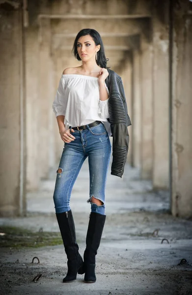 Portrait of beautiful sexy young woman with modern outfit, leather jacket, jeans, white blouse and black boots, in urban background. Attractive young brunette with long hair and blue eyes posing. — Stock Photo, Image