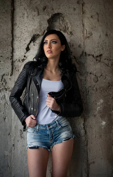 Portret van mooie sexy jonge vrouw met jeans, witte onderhemdje zwart lederen jas, in de stedelijke achtergrond. Aantrekkelijke brunette met perfecte lange benen op hoge hakken zilveren sandalen poseren mode. — Stockfoto