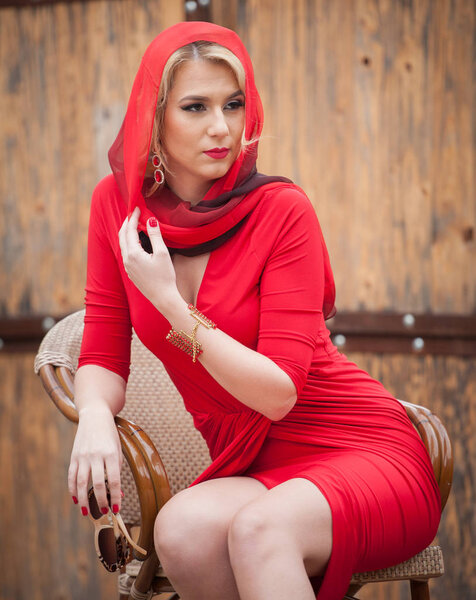 Fashionable attractive blonde woman in red dress sitting on chair. Beautiful elegant woman  with red scarf posing in elegant scenery .Sensual gorgeous young woman on high heels in red outfit