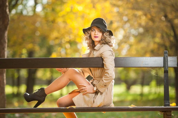 美丽的金发女子与奶油大衣 长腿和黑色的帽子在秋季的场景 一个非常美丽 优雅和感性的女人的肖像 卷曲的头发和性感的腿摆在秋季公园 — 图库照片