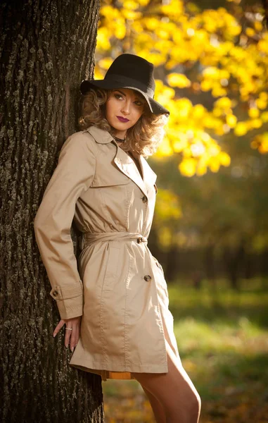 Beautiful blonde woman with cream coat, long legs and black hat in a autumn scene.  Portrait of a very beautiful, elegant and sensual woman with curly hair and sexy legs  posing in autumn park.