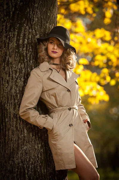 Beautiful blonde woman with cream coat, long legs and black hat in a autumn scene.  Portrait of a very beautiful, elegant and sensual woman with curly hair and sexy legs  posing in autumn park.