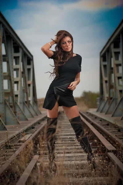 Atractiva mujer con vestido negro corto y botas de cuero largas de pie sobre los rieles con puente en el fondo. Moda chica sexy, en el puente posando en botas largas negras en tacones altos —  Fotos de Stock