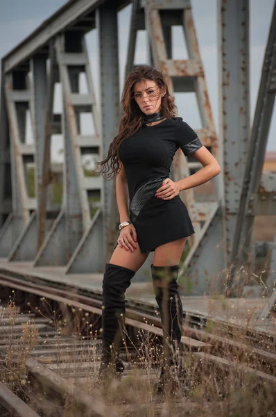 Jolie femme avec robe courte noire et de longues bottes en cuir debout sur les rails avec pont en arrière-plan. Mode fille sexy, sur le pont posant en bottes longues noires sur des talons hauts — Photo