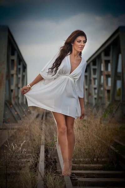 Atrakcyjna kobieta z krótką białą sukienkę i długie włosy, stojąc na szynach z bridge w tle. Moda sexy dziewczyna z sexy ciało i długie nogi na moście pozowanie w białej sukni — Zdjęcie stockowe