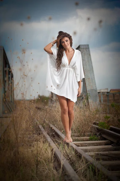 Mulher atraente com vestido branco curto e cabelos longos em pé sobre os trilhos com ponte no fundo. Menina sexy moda com corpo sexy e pernas longas na ponte posando em vestido branco — Fotografia de Stock
