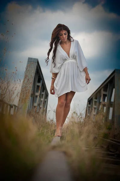 Mulher atraente com vestido branco curto e cabelos longos em pé sobre os trilhos com ponte no fundo. Menina sexy moda com corpo sexy e pernas longas na ponte posando em vestido branco — Fotografia de Stock