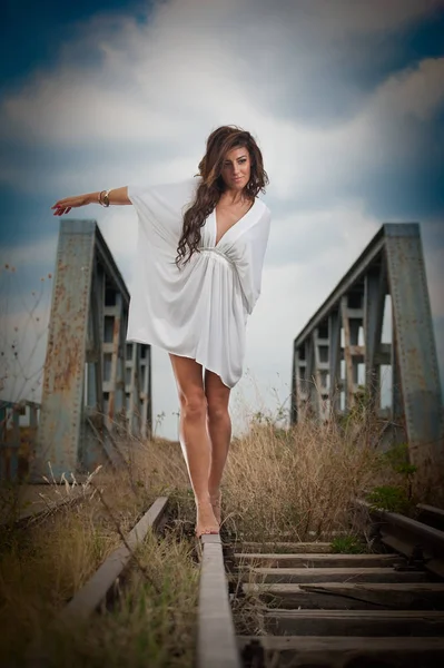 Atraktivní žena s krátké bílé šaty a dlouhé vlasy, stojící na kolejích s mostem v pozadí. Módní sexy dívka s sexy tělo a dlouhé nohy na mostě pózuje v bílých šatech — Stock fotografie