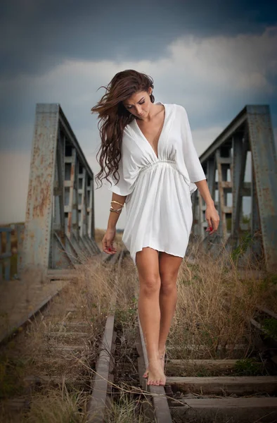 Atractiva mujer con vestido blanco corto y pelo largo de pie sobre los rieles con puente en el fondo. Moda chica sexy con cuerpo sexy y piernas largas en el puente posando en vestido blanco —  Fotos de Stock