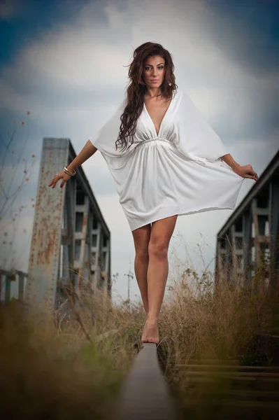 Attraktiv kvinne med kort hvit kjole og langt hår stående på skinnene med bro i bakgrunnen. Motesexy jente med sexy kropp og lange bein på broen poserer i hvit kjole – stockfoto
