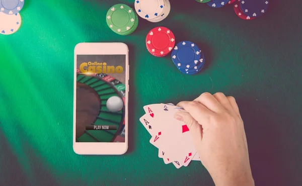 smartphone with online casino website on screen, chips and cards over green poker table, male hand holding cards. Gambling concept